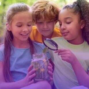 three kids are gathered around a glass jar filled with plants and flowers